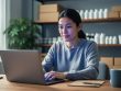 Small business owner typing on a laptop at a neat studio desk with soft window light, sharp focus on hands and face, and blurred shelves of products, packing boxes, and a plant in the background; laptop screen content is intentionally indistinct with no readable text.