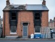 Street-level view of a fire-damaged UK terraced house exterior with soot-stained brickwork and repaired roof sections under natural daylight.